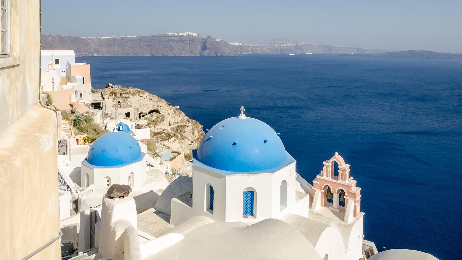 Witte kerken met blauwe koepels en een roze klokkentoren in Oia boven de caldera